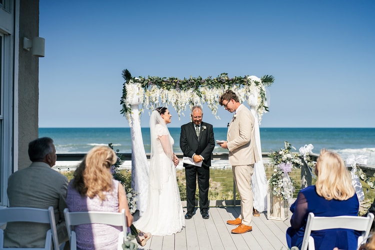 Bride and groom exchanging vows on the beachfront deck; groom reads from a card while the bride smiles, the blue ocean visible behind them.