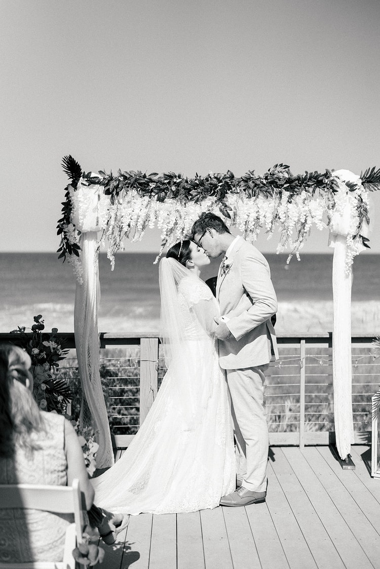 Black-and-white image of bride and groom sharing their first kiss beneath the floral ceremony arch as guests watch.
