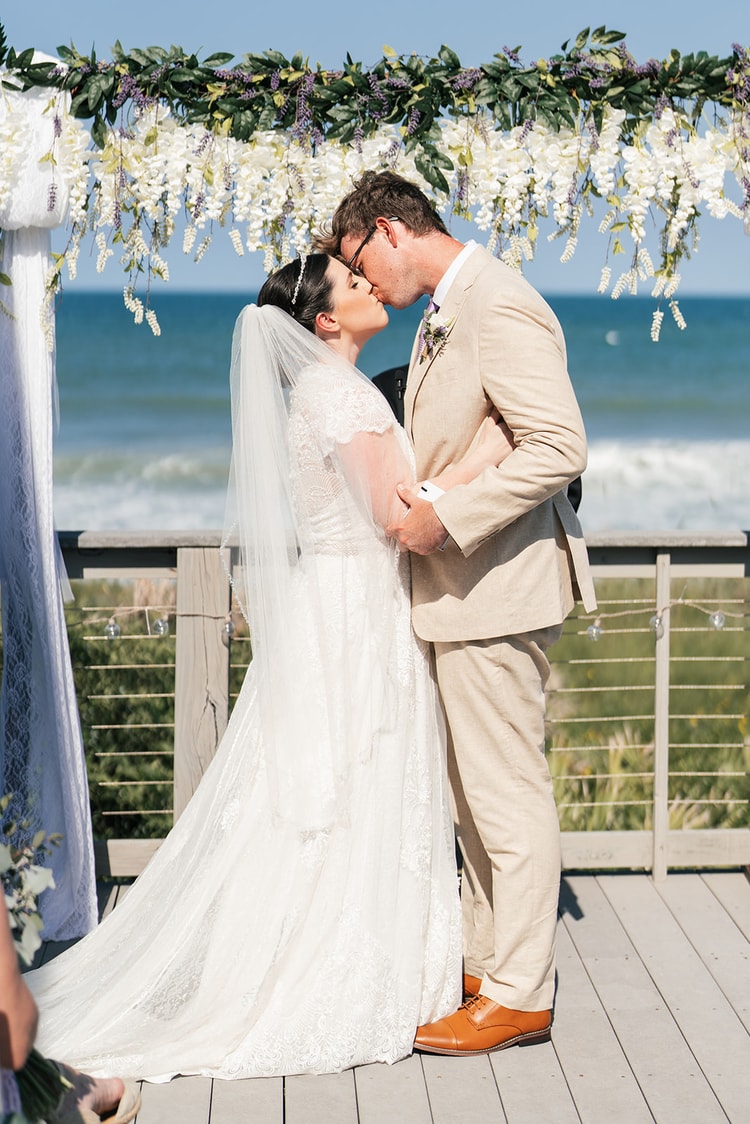 Bride and groom sharing their first kiss under a white wisteria arch with the ocean sparkling behind them.