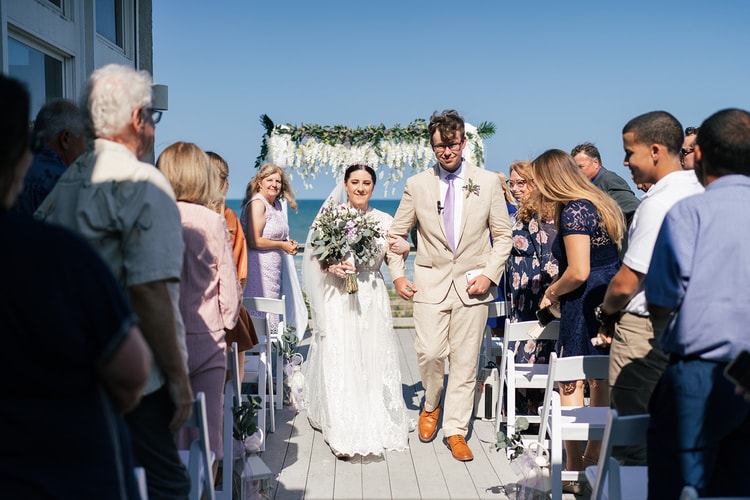 Bride and groom walking hand in hand down the aisle after the ceremony, beaming, with the floral arch and ocean behind them and cheering guests on either side.
