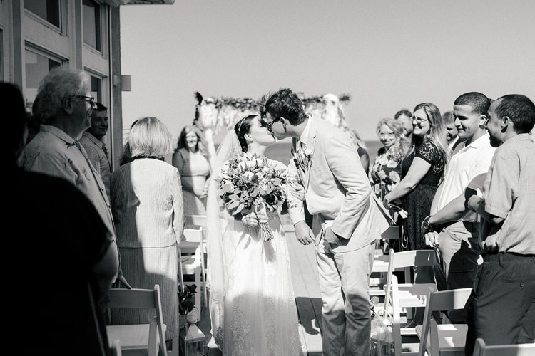 Black-and-white recessional moment: bride and groom steal a kiss mid-aisle as standing guests cheer around them.