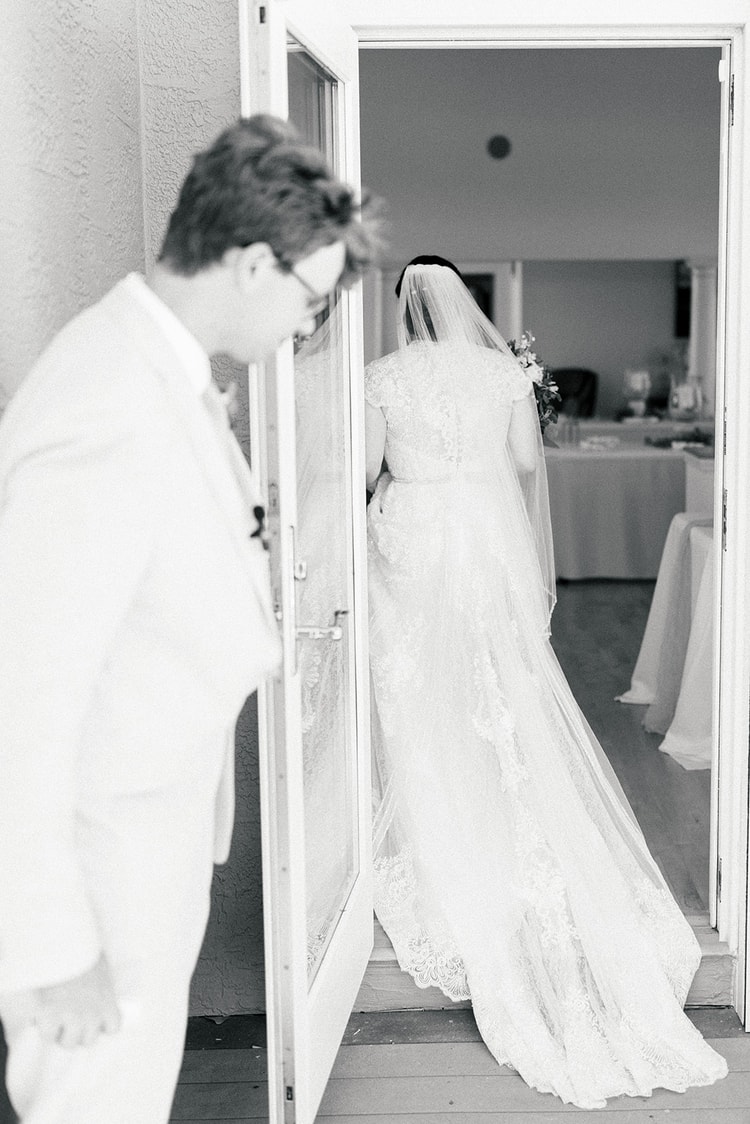 Black-and-white image of the groom holding a door open as the bride, veil trailing, steps inside just after the ceremony.