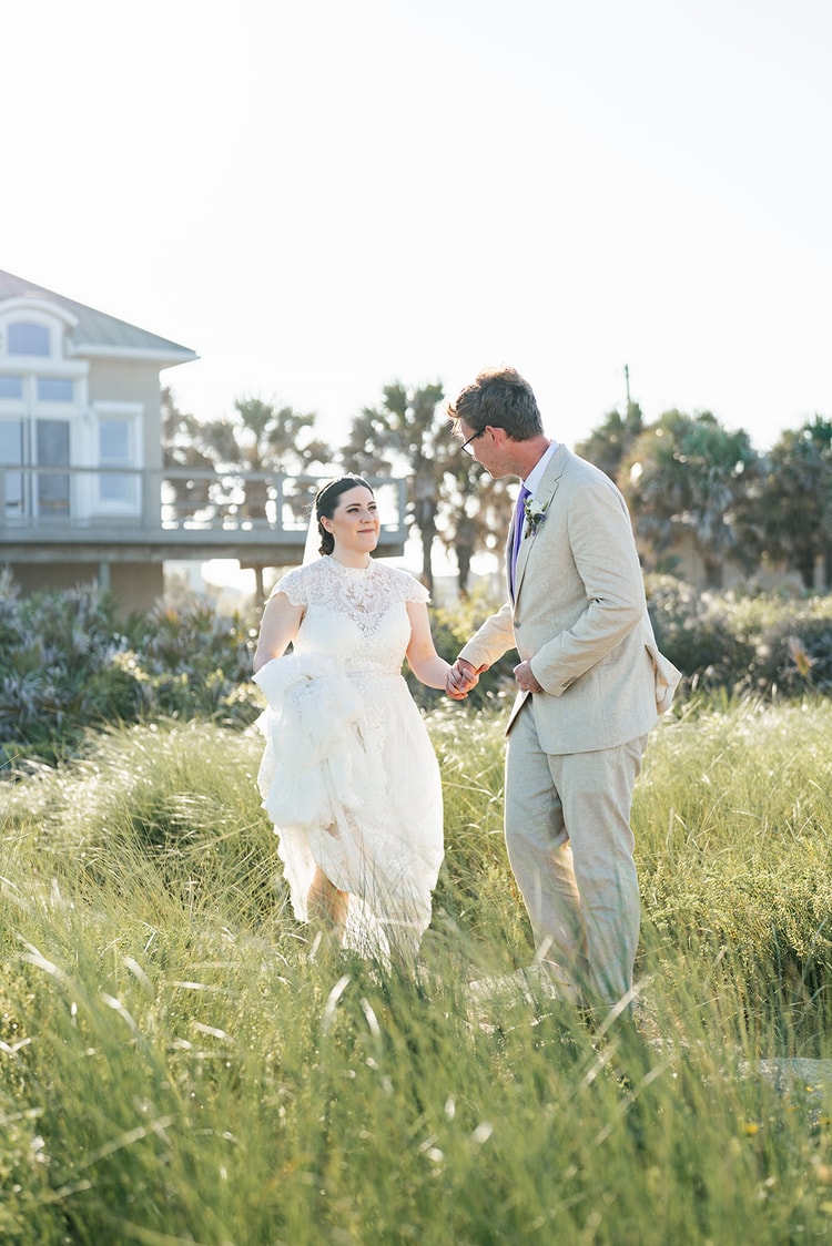 Bride and groom face each other holding hands while walking through tall sea grass near the beach at golden hour