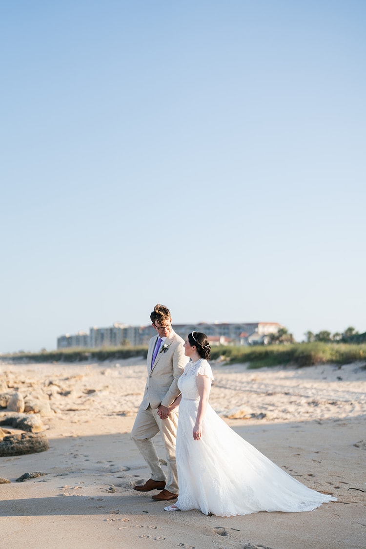 Couple walks hand in hand on a sandy beach at golden hour, the bride's train trailing behind her