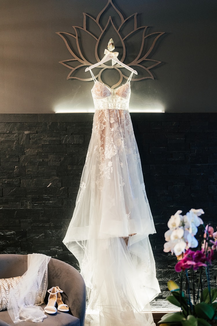 Bridal gown with delicate lace applique hanging from a white hanger in front of a dark stone wall, heels placed on the chair below.