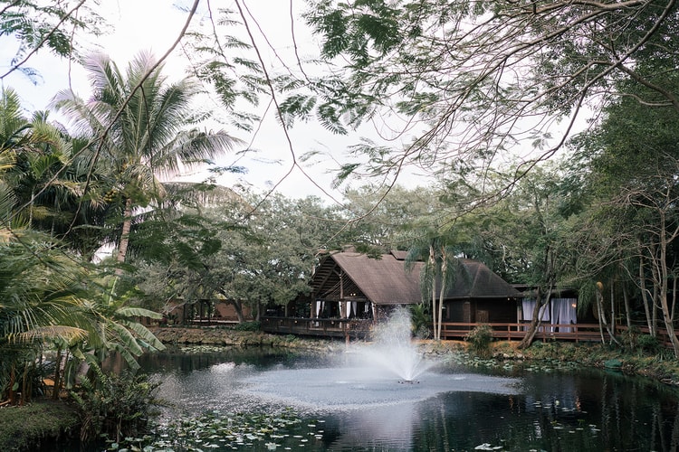 Rustic venue building reflected in a still pond surrounded by palm trees and lush greenery, a fountain rippling at the center.