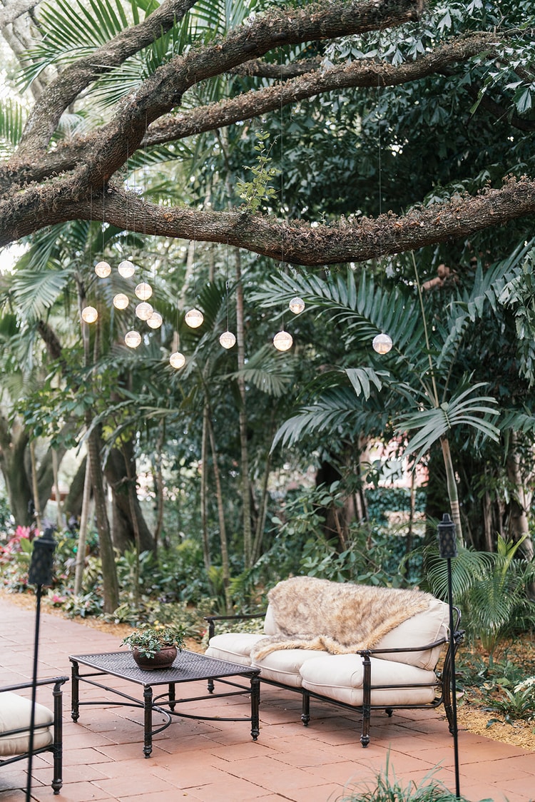 Outdoor cocktail lounge area with a loveseat and coffee table beneath hanging globe lights and sweeping tree branches.