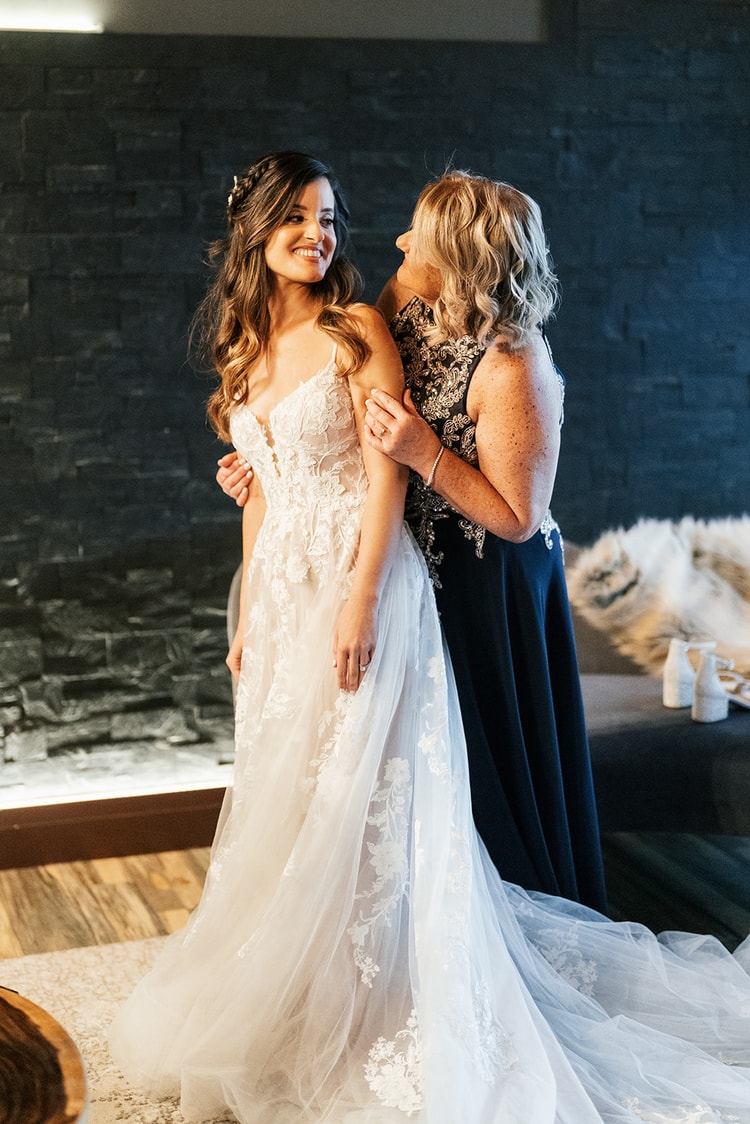 Bride in her lace wedding gown smiling as a woman in a navy dress adjusts her shoulders during getting-ready preparations.