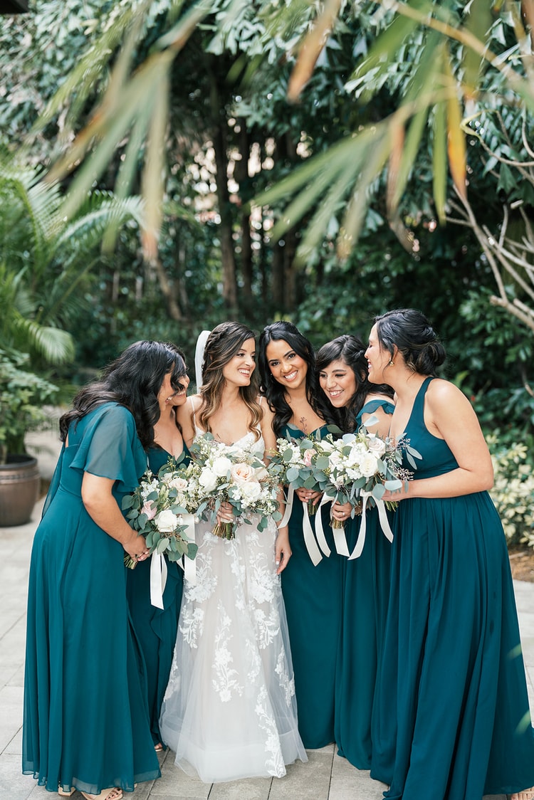 Bride surrounded by four bridesmaids in teal gowns, all laughing and leaning in together beneath tropical palms.