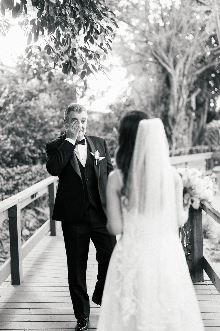 Black and white portrait of a father wiping tears from his eyes on a wooden bridge as he sees his daughter in her wedding gown for the first time.