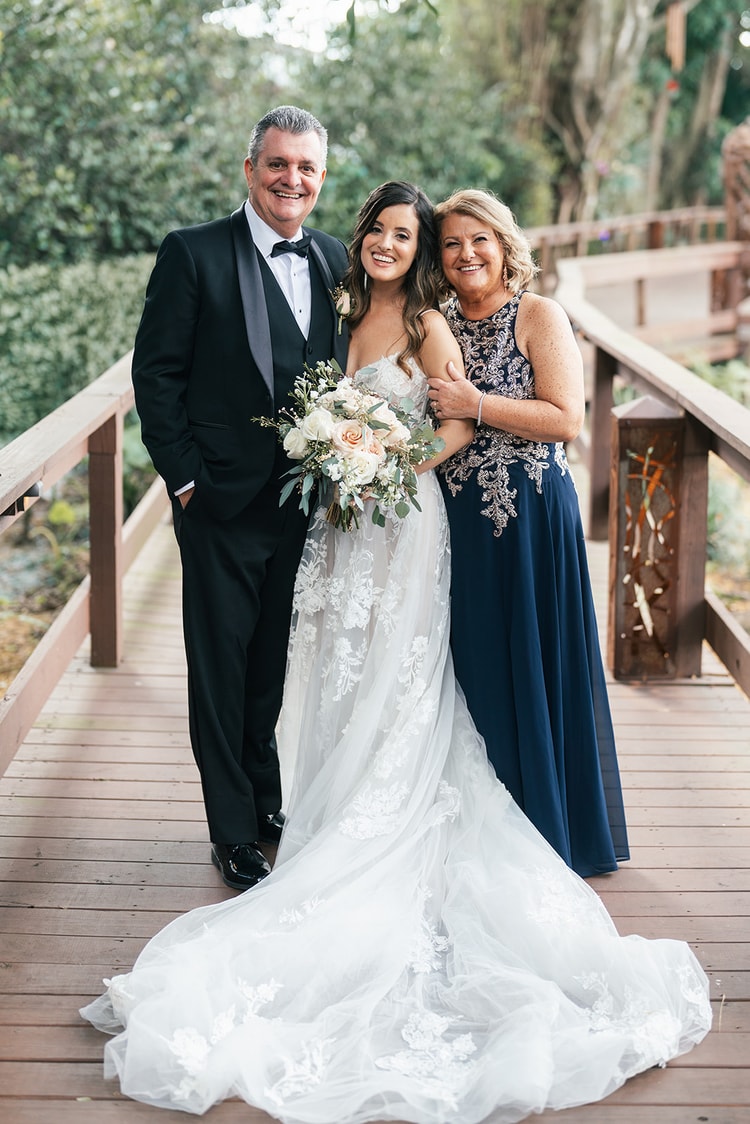 Bride standing between her parents on a wooden bridge, all three smiling warmly before the ceremony.