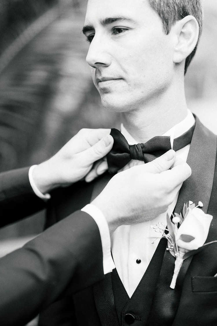 Black and white close-up of a groomsman adjusting the groom's bow tie, a boutonniere pinned to the lapel.