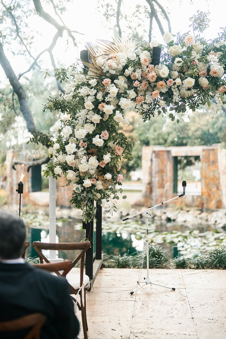 Ceremony arch towering with lush blush and ivory roses, eucalyptus, and gold-tipped palm leaves beside a reflective pond.