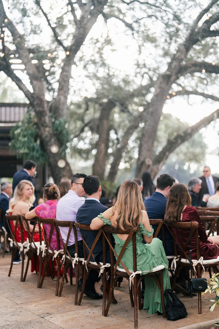 Guests seated in cross-back chairs beneath sprawling oak trees at an outdoor wedding ceremony, twinkling lights overhead.
