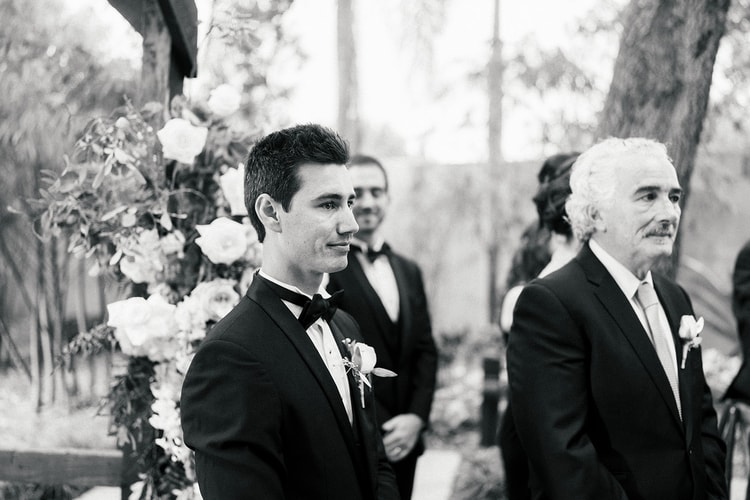 Black and white ceremony moment showing the groom at the altar, looking down the aisle as guests and family stand nearby.
