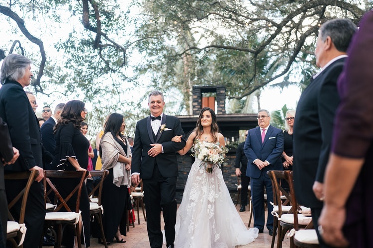 Bride walking down the aisle on her father's arm, guests rising and smiling on both sides, stone archways and trees in the background.