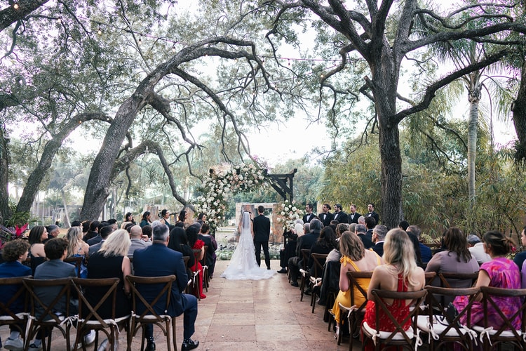 Wide view of the outdoor wedding ceremony with the couple at the flower-draped altar, guests filling the seated area beneath ancient oak trees.
