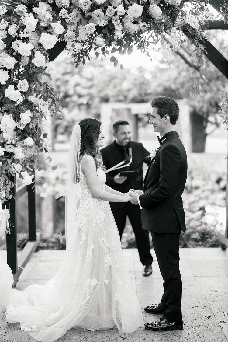 Black and white vows moment: bride and groom hold hands beneath a floral arch as the officiant reads behind them.