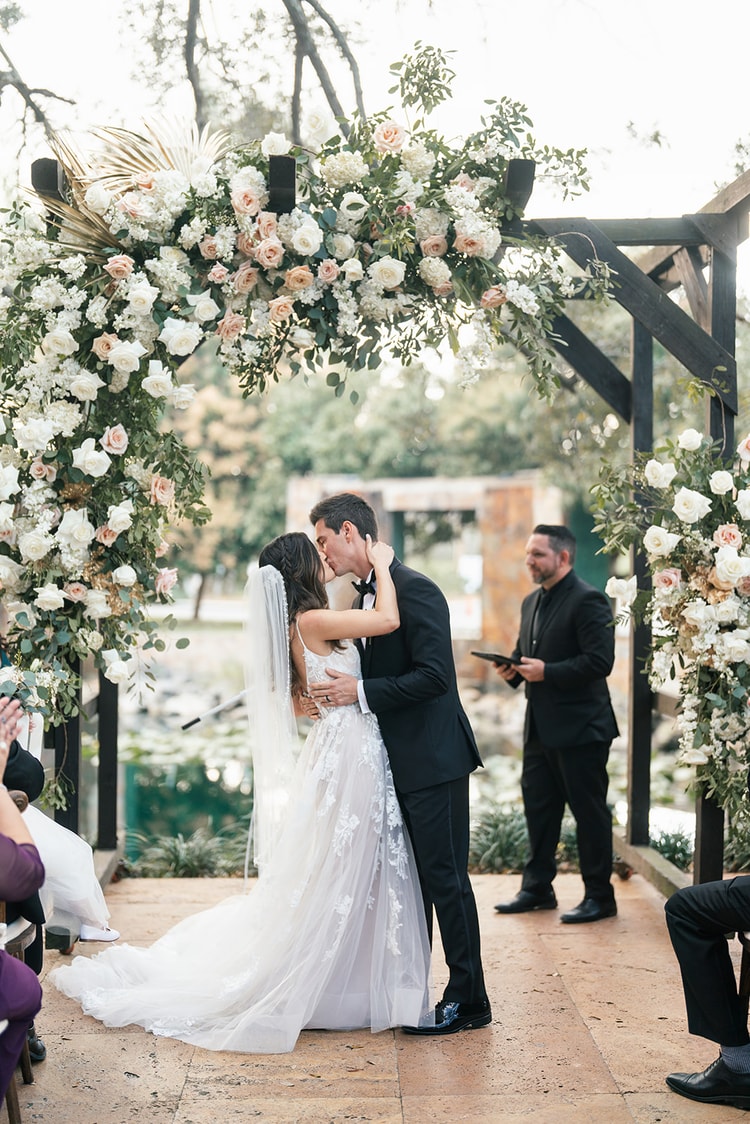 Bride and groom share their first kiss under a lush floral arch of blush and white roses, guests applauding around them.