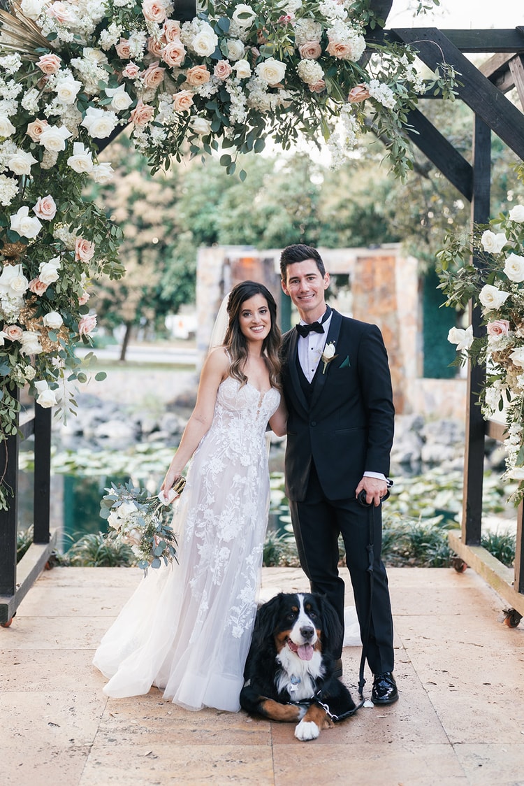 Newlyweds pose together beneath the floral ceremony arch with their Bernese Mountain Dog sitting at their feet beside the pond.