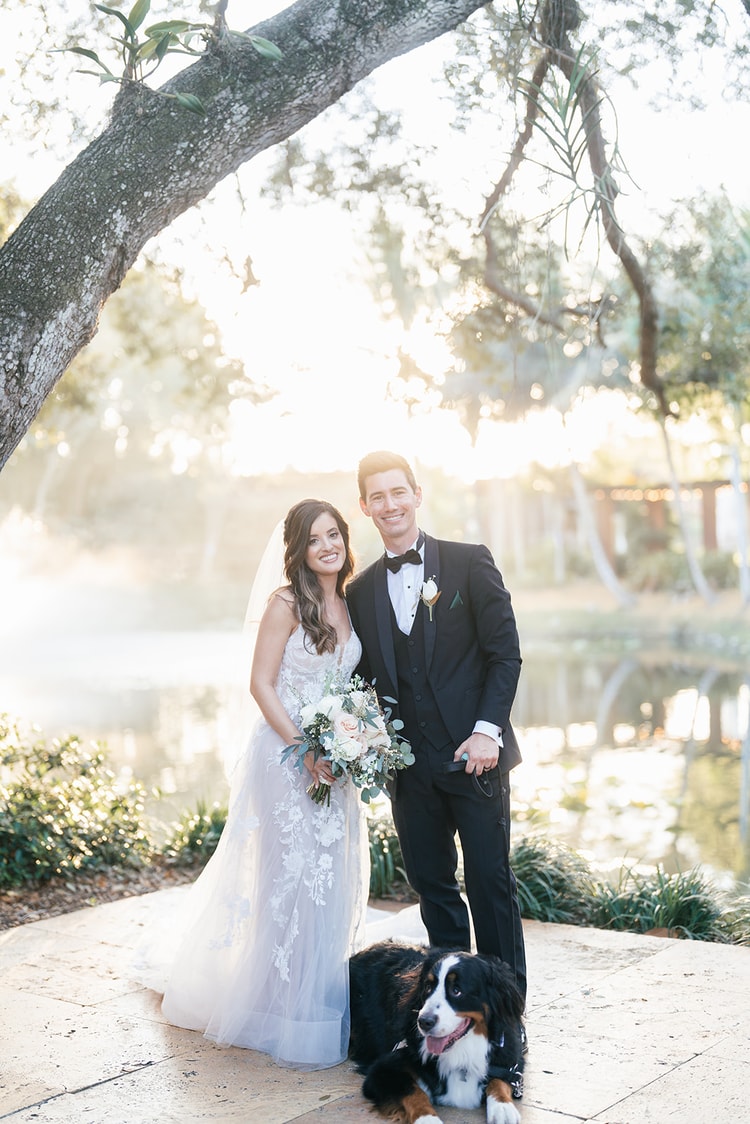 Bride and groom portrait beside a misty pond at sunset, their Bernese Mountain Dog resting at their feet beneath Spanish moss.