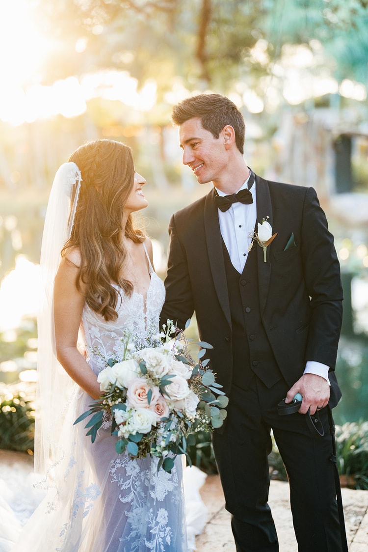 Bride and groom gazing at each other during portrait time in warm golden-hour light, bouquet of soft blush roses between them.