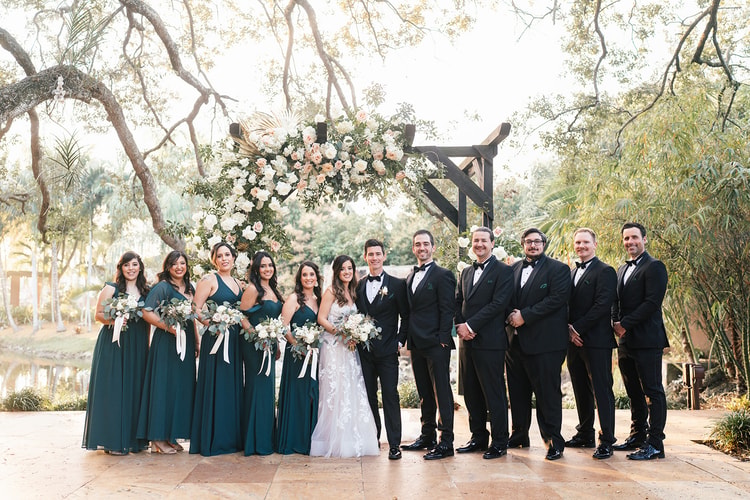 Wedding party portrait outdoors with the couple kissing at center while bridesmaids and groomsmen laugh and react around them.