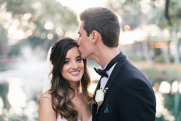 Bride and groom walking together along a stone wall path, framed by a palm frond overhead and a stone archway behind.