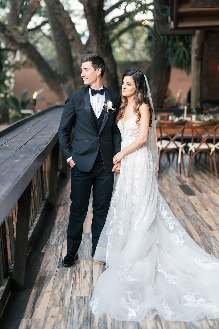 Bride resting her head on the groom's shoulder as they stand on a wooden bridge at dusk, candlelit reception tables behind them.