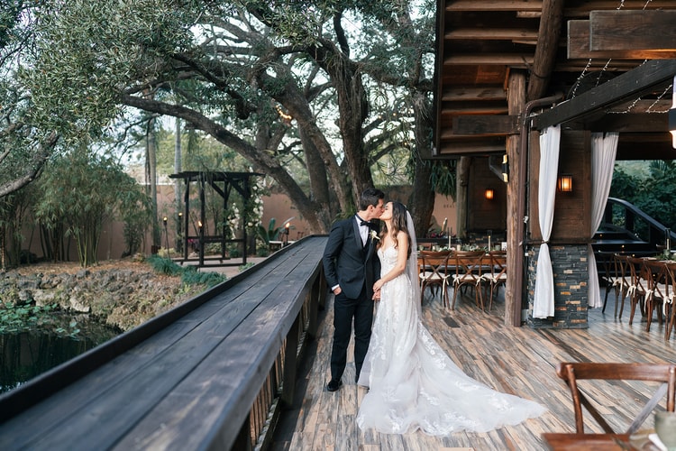 Couple sharing a kiss on a wraparound deck as the reception area glows with candlelight and hanging lights in the trees behind them.