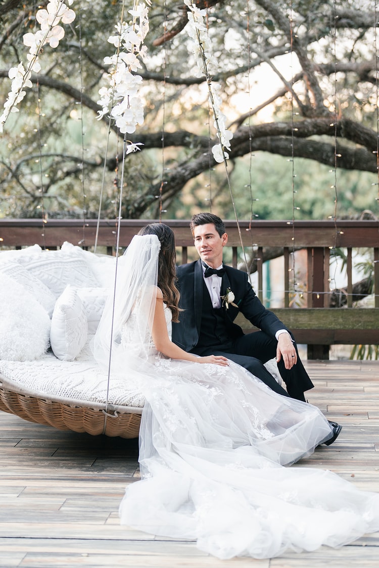 Bride and groom seated on an outdoor hanging daybed, relaxed and smiling at each other beneath floral and twinkle light decorations.