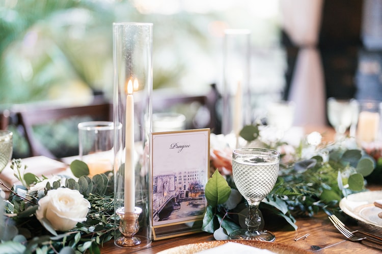 Reception tablescape detail featuring a travel-themed table number card reading Prague, hurricane candle, cut-crystal glassware, and white roses.