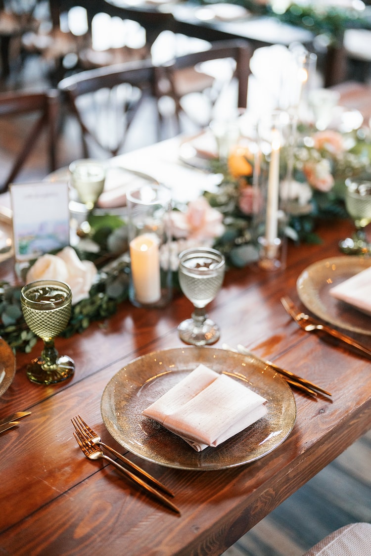 Reception place setting close-up with a gold charger, blush napkin, gold utensils, and a textured green goblet on a wooden farm table.