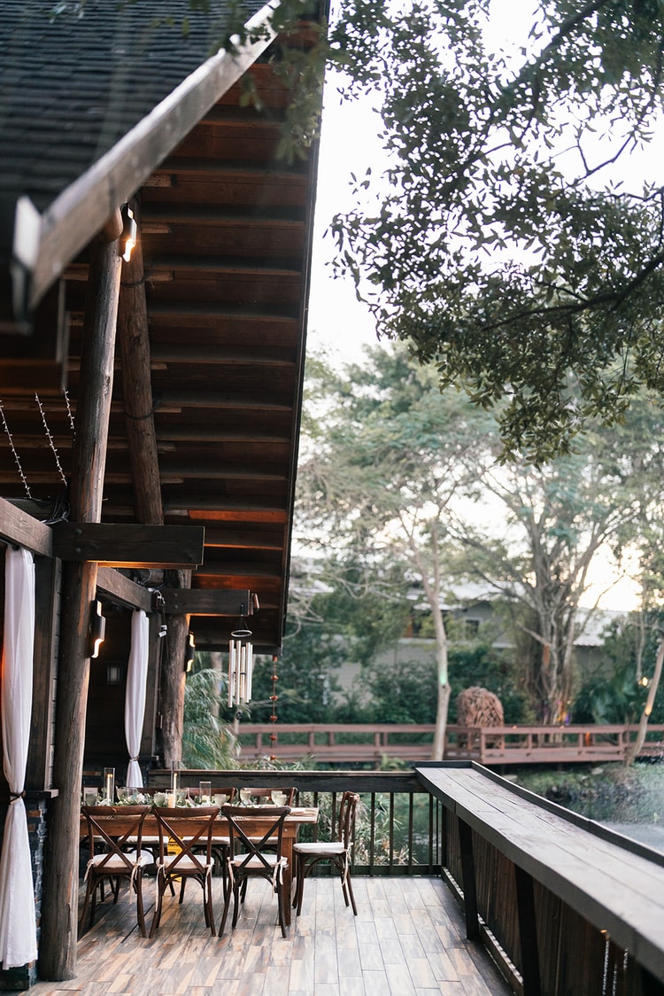 Rustic outdoor reception space viewed from the side, a long wooden farm table set beneath a timber-framed pavilion overlooking a pond.