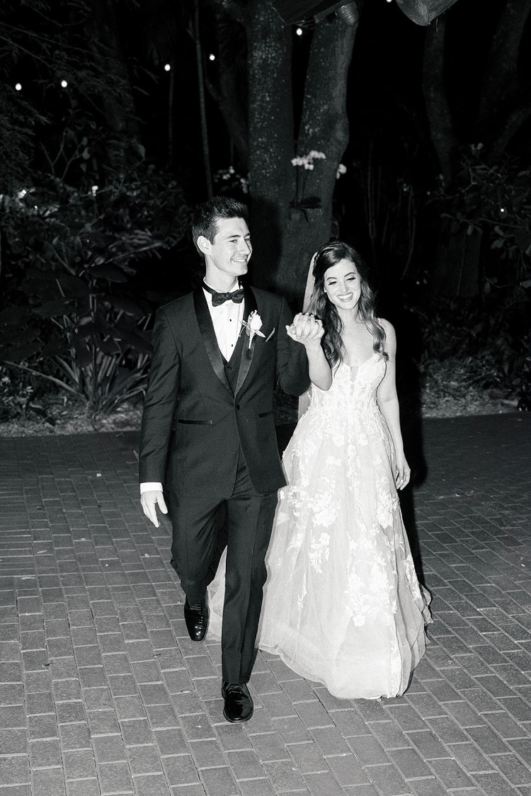 Bride and groom walking hand in hand at night beneath garden trees, laughing as they make their reception entrance.
