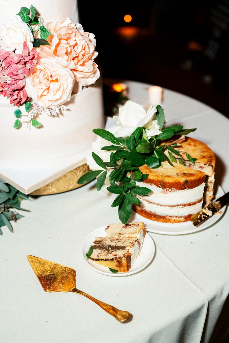 Flower girl in white dress concentrating on her cake slice at the reception table, guests dancing in the background.