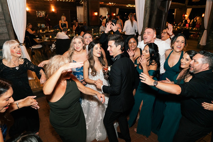 Couple and guests celebrating on the reception dance floor under string lights, bride and groom surrounded by cheering family and friends.
