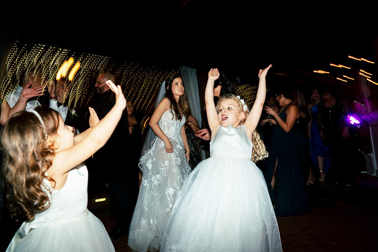 Bride dancing joyfully on the reception floor under a cascade of string lights as flower girls in white dresses throw their arms up beside her.