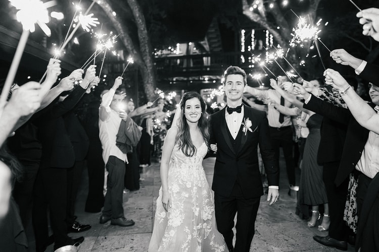 Couple framed beneath a flowering garden arch glowing with lights at night, holding hands as they walk through the luminous tunnel.