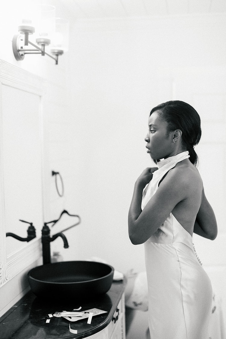 Bride in a halter wedding gown fastening her collar in front of a bathroom mirror, black-and-white