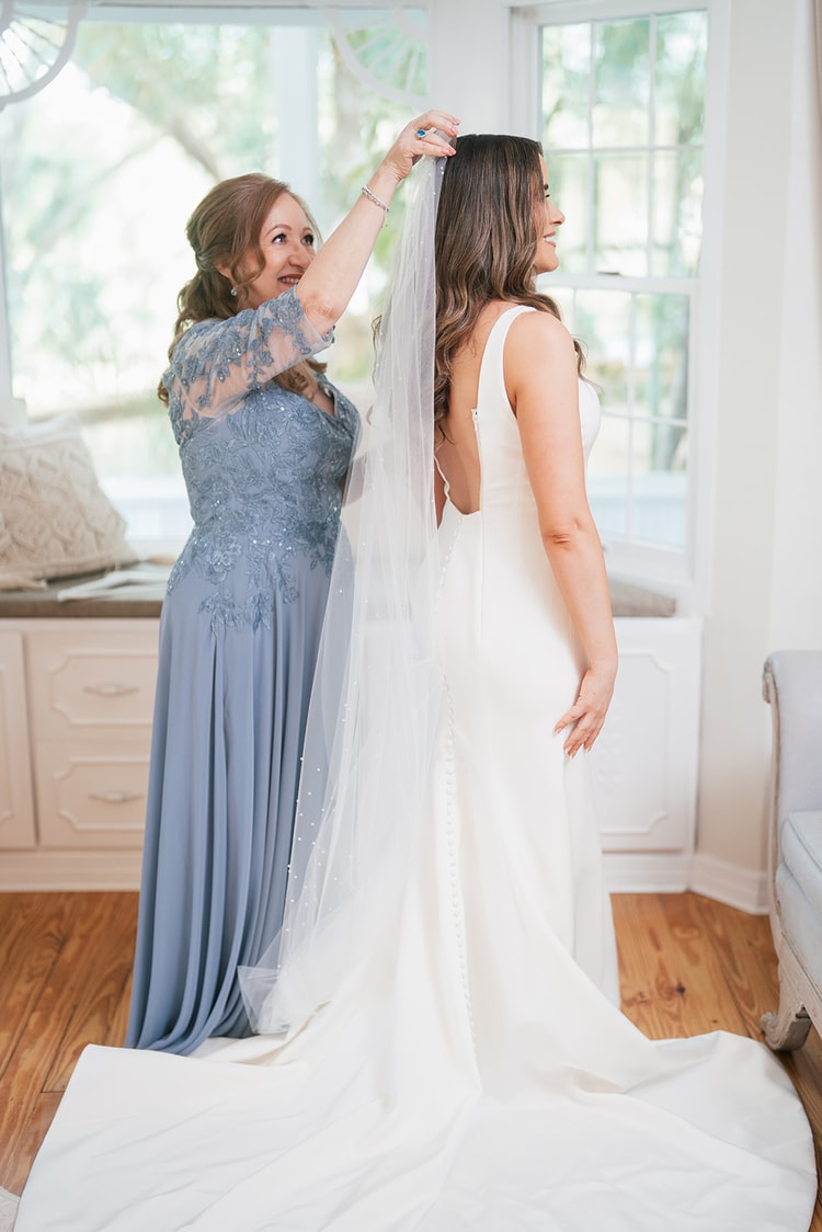 Mother in blue lace gown placing the pearl-dotted veil onto the bride's hair in a bright room
