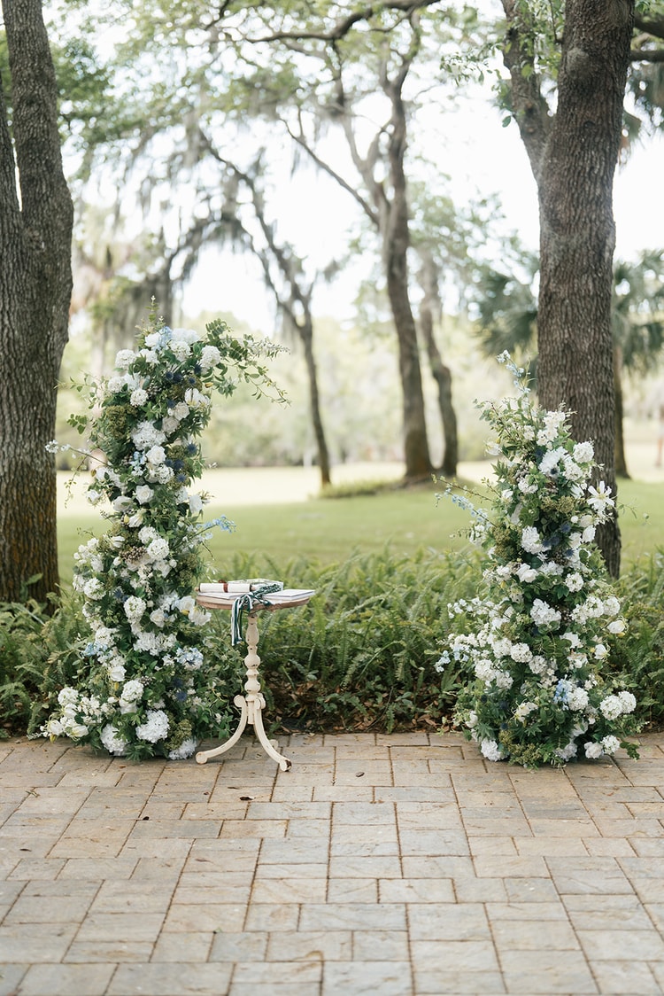 Pair of tall floral columns covered in white hydrangeas, blue delphinium and greenery flank a ceremony pedestal table on a brick path, wedding details