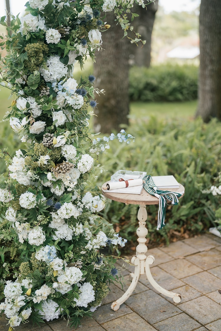 Close detail of a floral ceremony column with white hydrangeas and blue thistle beside a pedestal table holding vow scrolls and a braided cord, wedding details