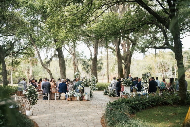 Wide view of guests seated at an outdoor ceremony beneath sweeping oak trees draped in moss, flanked by floral arrangements, wedding photography