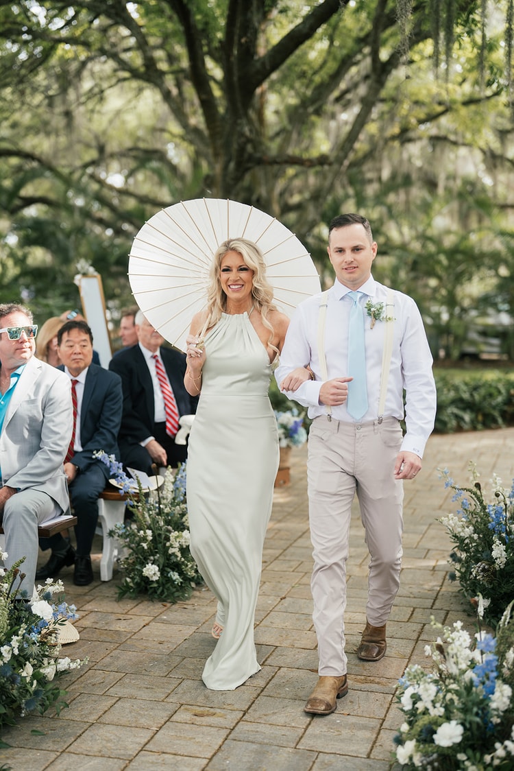 Woman in a sage gown carries a white parasol while escorted down the aisle under oak trees as guests look on, wedding photography