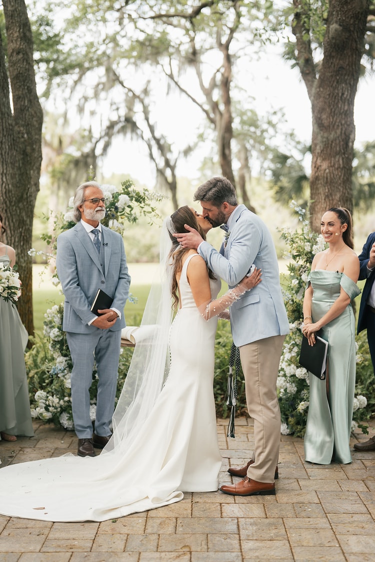 Couple share their first kiss at a moss-draped outdoor altar, officiant and wedding party smiling around them, wedding photography