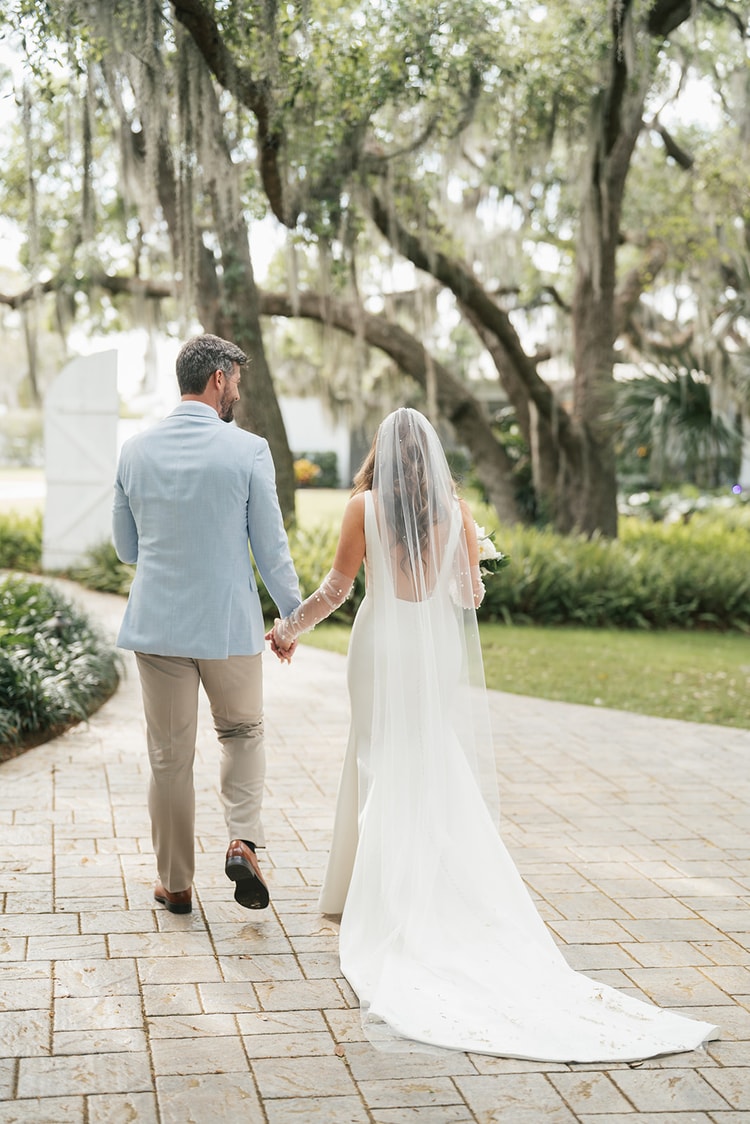 Couple walk hand in hand away from camera along a brick path beneath arching moss-draped oaks, bride's pearl veil trailing behind, wedding photography