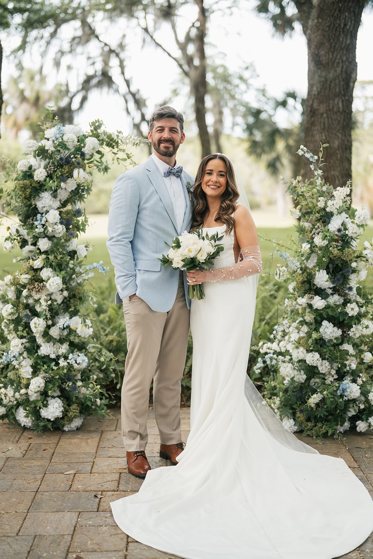 Bride and groom stand together smiling before a lush blue and white floral arch on a brick patio beneath Spanish moss-draped trees