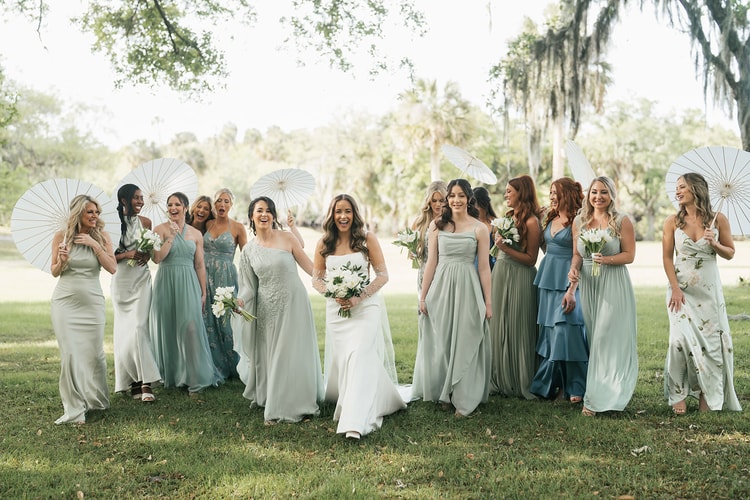 Bride walks with a large group of bridesmaids in mixed sage, blue and floral gowns across a lawn, several holding white parasols, wedding photography