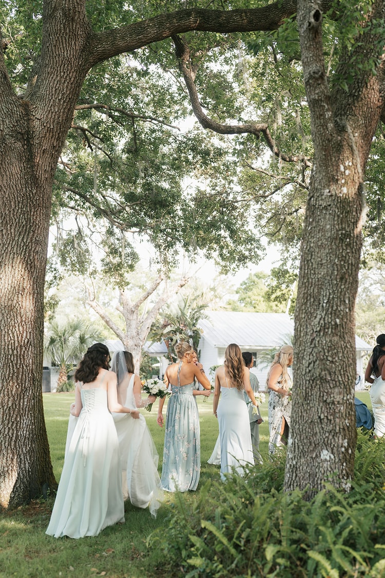 Bridal party gathers on a grassy lawn between two large oak trees, bride and bridesmaids in soft sage and blue gowns, wedding photography